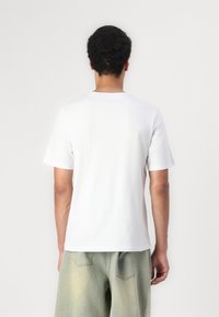 Hombre con cabello rizado y corto que lleva una camiseta blanca de manga corta lisa y pantalones cortos de mezclilla verde claro, visto de espaldas sobre un fondo liso.