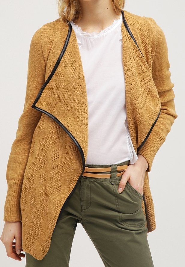 Mujer con cárdigan tejido de color mostaza con textura y abertura frontal, con ribete negro, blusa blanca con volantes, pantalones verde oliva y cinturón marrón claro.