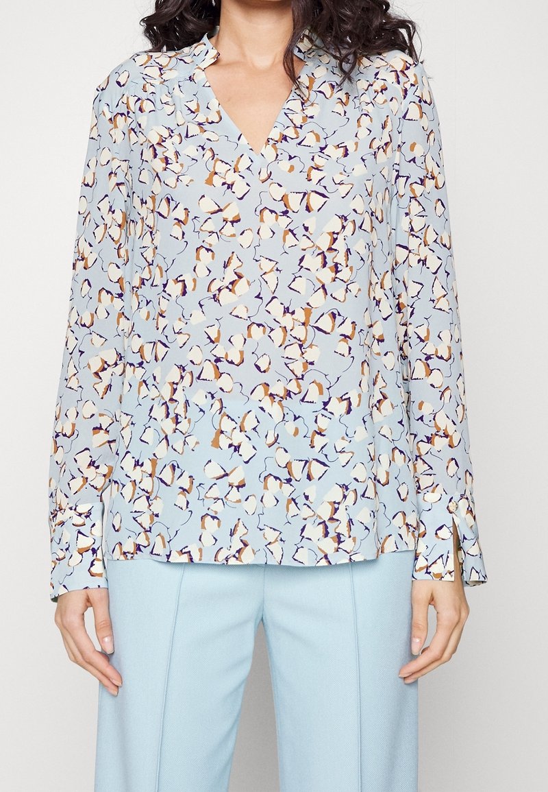 Bluse in Hellblau mit einem floralen Muster in Orange und Lila. Sie hat einen V-Ausschnitt, lange Ärmel und einen fließenden Stoff. 