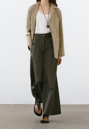 Vrouw met een beige blazer over een wit topje, losse, donkergroene broek met hoge taille, zwarte sandalen en een zwarte hangerketting, lopend naar voren.