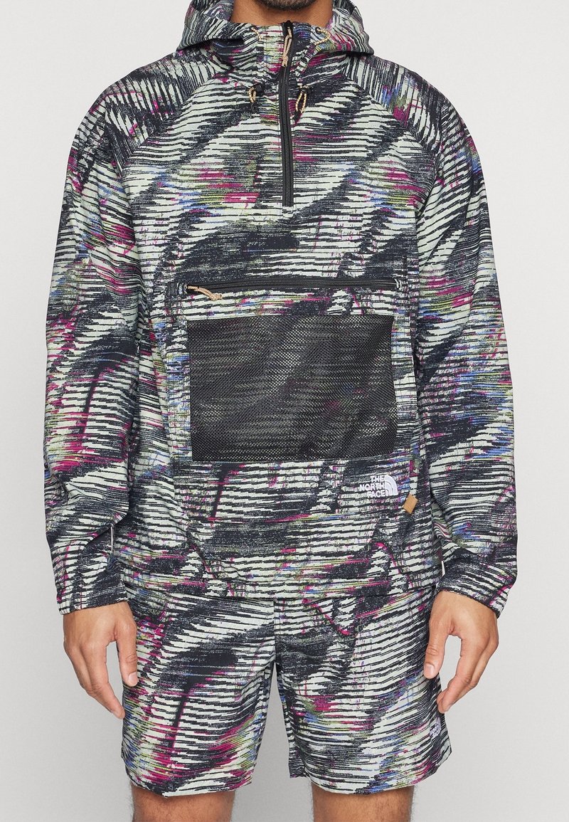 Homme portant un pull à capuche avec un motif abstrait multicolore, une poche frontale en mesh noir et un short assorti.