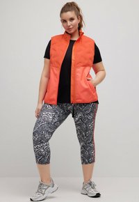 Orange vadderad väst med hög krage, i kombination med svart skjorta och mönstrade grå leggings, har en slim fit och sidofickor.