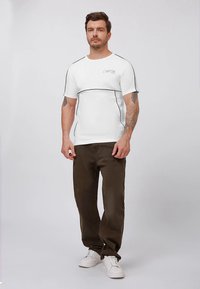 Bela kratka majica s črnimi obrobami in logotipom, kombinirana z olivno zelenimi hlačami in belimi supergami, na enobarvnem ozadju.