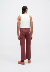 Homem com cabelo trançado, vestindo uma blusa branca sem mangas, calças bordô com bolsos traseiros e sapatos laranja, de pé de costas contra um fundo branco.