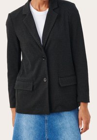 Blazer a quadri neri con revers a toppa, due tasche frontali e chiusura con un bottone. Indossato sopra una t-shirt bianca, la trama è sottile.