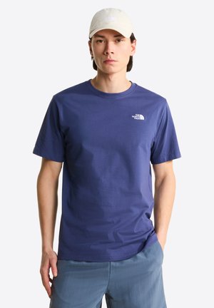 Młody mężczyzna w kremowej czapce, niebieskiej koszulce The North Face i jasnoniebieskich spodniach, stojący z jedną dłonią w kieszeni na jednolitym tle.