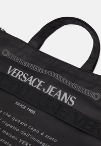 Sac fourre-tout en tissu noir avec le texte "VERSACE JEANS" en gris et une section "COUTURE" en relief. Comprend un graphique de chaîne et des poignées arrondies.