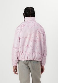 Light pink jacket with a textured finish, featuring a wavy pink pattern, elastic cuffs, and a high collar. Paired with light denim jeans.