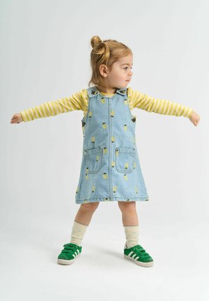 Džínové šaty s žltými potlačami ananásov, so sieťovanými prednými vreckami, skombinované so žltým pruhovaným tričkom s dlhým rukávom a zelenými teniskami na suchý zips.