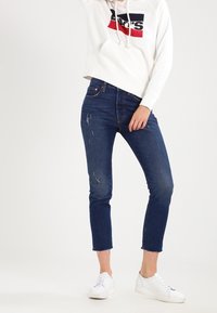 Personne portant un jean skinny raccourci bleu foncé avec des détails usés, des baskets blanches et un sweat à capuche blanc Levi's avec logo.