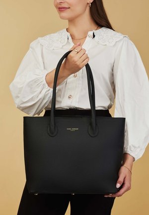 Zwarte lederen totebag met gestructureerd oppervlak, twee stevige handgrepen en een gouden logo-detail aan de voorkant. Eenvoudig, rechthoekig ontwerp.