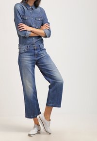 Blå denimskjorta med knäppning framtill och två bröstfickor, kombinerad med ljusblå vida jeans och vita sneakers. Slät textur hela vägen.