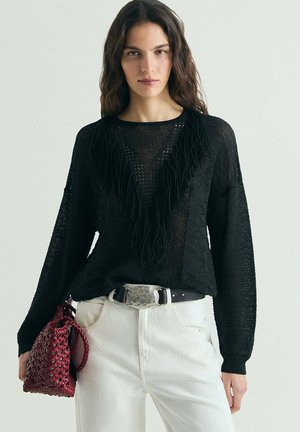 Mujer con un suéter negro con flecos metido en pantalones blancos con un cinturón con una gran hebilla plateada, sosteniendo un bolso rojo texturizado.