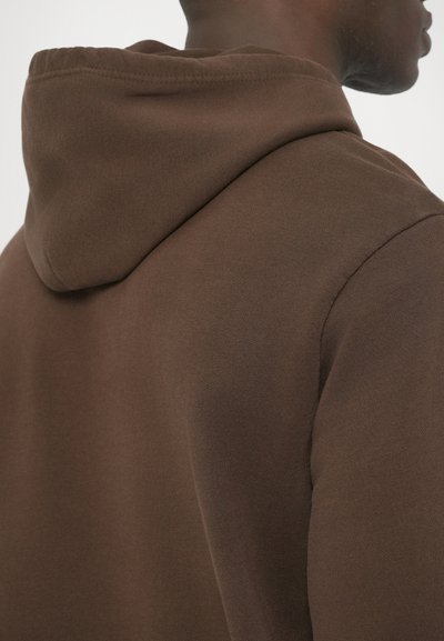 Barna pamut kapucnis pulóver puha tapintással, kerekített kapucnival és minimális varrási részletekkel. Oldalról látható, kiemelve a váll területét.