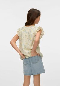 Blusa leggera gialla a fantasia floreale con maniche arricciate, abbinata a una gonna di jeans. La blusa presenta una vestibilità comoda e un tessuto testurizzato.