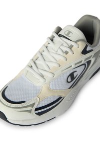 Zapatilla deportiva blanca y beige con parte superior de malla, interior gris, acentos en negro y gris, cordones blancos y un logo en el lado y la lengüeta.