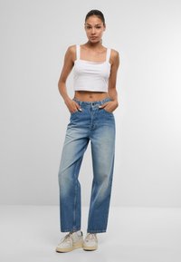 Balta īsā topiņa ar kvadrātveida izgriezumu, kopā ar gaiši zilām platām biksēm. Valkāta ar bēšiem sporta apaviem, rokas kabatās.