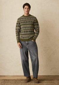 Egy férfi zöld mintás pulcsit, laza szürke farmert és barna cipőt visel, egy szőtt szőnyegen áll egy bézs fal előtt.