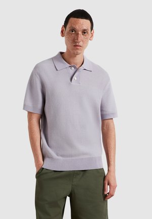 Jonge man met kort donker haar en sproeten, gekleed in een lichtpaarse, gestructureerde korte mouw polo en een groene broek, met de handen in de zakken.