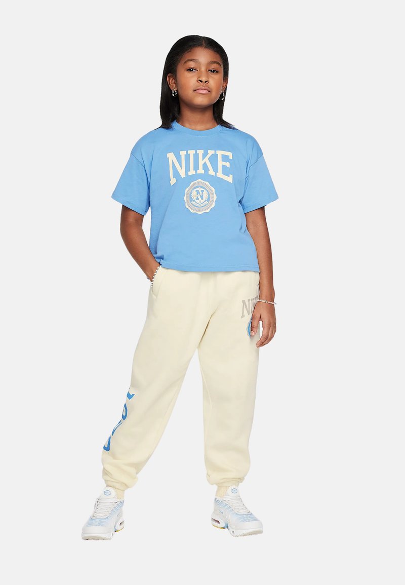 Camiseta de algodón azul con el logo "NIKE", combinada con pantalones de chándal crema con acentos azules y zapatillas ligeras con suela tipo burbuja.