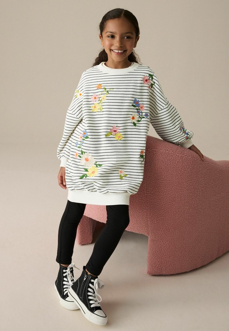 Pige smilende, iført en hvid stribet sweaterkjole med farverige blomster, sorte leggings og sorte høje sneakers, læner sig op ad en lyserød tekstureret stol.