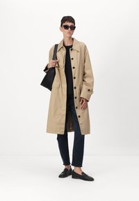 Beige bomull trench coat med store knapper, sidelommer og en avslappet passform, sammen med svart topp, mørke jeans og svarte sko.