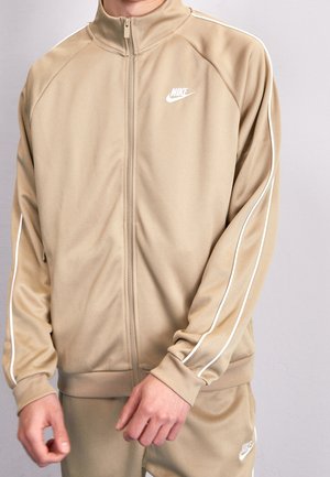 Beżowy dres Nike z białymi lamówkami na rękawach, kurtka zapinana na zamek oraz pasujące spodnie, noszone przez osobę z rozluźnionymi rękami z przodu.