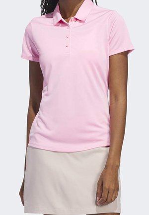 Mujer con una camisa polo de manga corta color rosa claro y falda beige, de pie contra un fondo liso y claro.