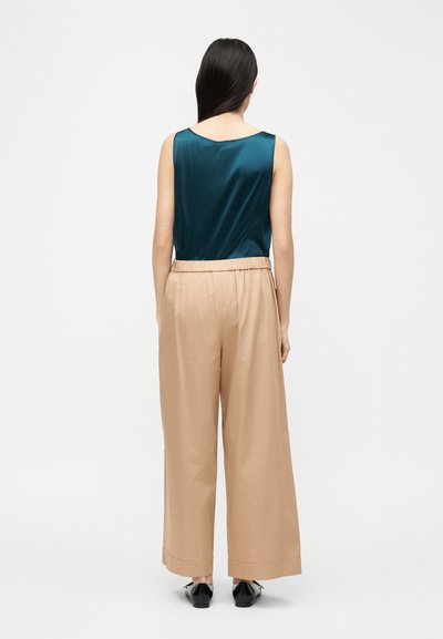 Γυναίκα με μακριά σκούρα μαλλιά, ντυμένη με τιρκουάζ μπλουζάκι χωρίς μανίκια, μπεζ παντελόνι με φαρδιά πόδια και μαύρα παπούτσια, στέκεται με την πλάτη προς τα εμπρός σε ένα απλό φόντο.