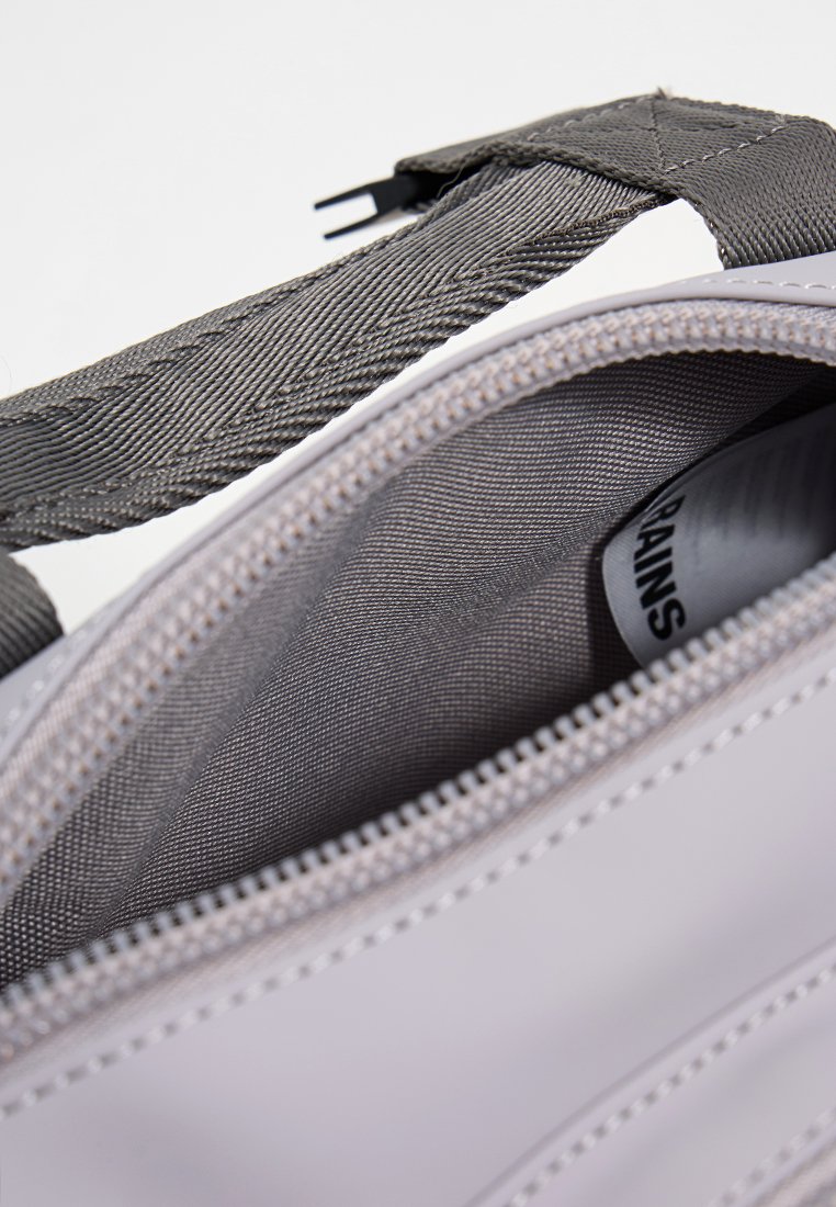 Grijze tas met een gestructureerde schouderband, witte rits en een glad, glanzend oppervlak. De binnenkant heeft een eenvoudige grijze voering.