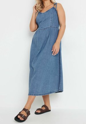 Kvinde iført en ærmeløs blå denim midi-kjole med bindebånd på skuldrene og brune sandaler med nitter, stående op ad en hvid væg.