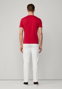 Rode aansluitende t-shirt, korte mouwen, achteraanzicht, gecombineerd met slanke witte broek en donkere sneakers. Eenvoudig ontwerp, geen zichtbare patronen of logo's.