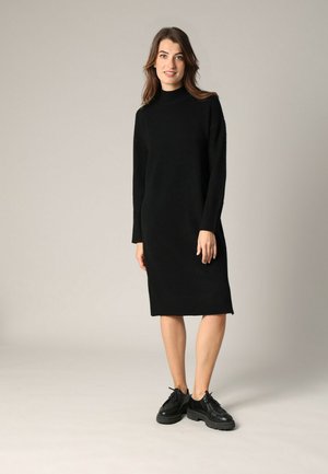 Femme debout portant une robe pull midi à manches longues noire et des chaussures à lacets noires à semelles épaisses sur un fond gris clair.