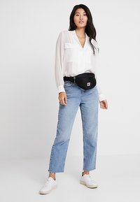 Donna che indossa una camicia bianca, jeans azzurri chiari, sneakers bianche e una borsa nera a vita, in piedi di fronte a uno sfondo bianco semplice.