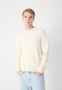 Suéter de punto acanalado en color crema con cuello redondo y mangas largas, combinado con jeans de mezclilla azul claro, con las manos en los bolsillos.