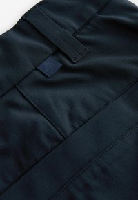 Marineblauer Stoff mit einer glatten Textur, der ein rechteckiges Etikett aufweist. Das Design beinhaltet dezente Nähdetails und einen flachen Taillenbund.