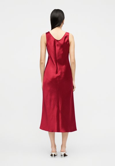 Γυναίκα με μακριά σκούρα μαλλιά φοράει αμάνικο, λαμπερό κόκκινο φόρεμα midi και λευκά mule με τακούνι, στέκεται μπροστά σε απλό λευκό φόντο.