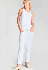Débardeur blanc côtelé avec texte imprimé, associé à un pantalon blanc à jambes droites. Le mannequin porte des mocassins beige. Design simple et épuré.
