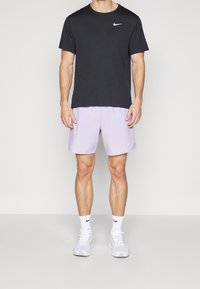 Camiseta negra de Nike de manga corta con un logo blanco, combinada con pantalones cortos púrpura claro y zapatillas blancas. El modelo está de pie contra un fondo liso.