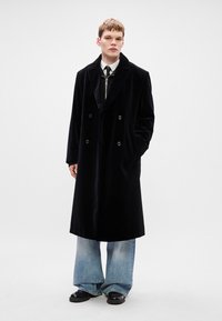 Μαύρο βελούδινο διπλής όψης παλτό με καπιτονέ πέτα, πλαϊνές τσέπες και μαύρα κουμπιά, φορεμένο πάνω από ανοιχτόχρωμο τζιν παντελόνι σε φαρδιά γραμμή.