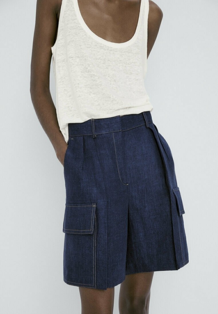 Short en jean bleu foncé taille haute avec de grandes poches avant, porté avec un débardeur blanc sans manches sur une personne debout.