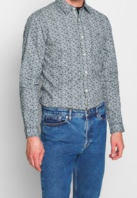 Man som bär en långärmad skjorta med blå blommönster, instoppad i högmidjade blå jeans, som står mot en enfärgad bakgrund.