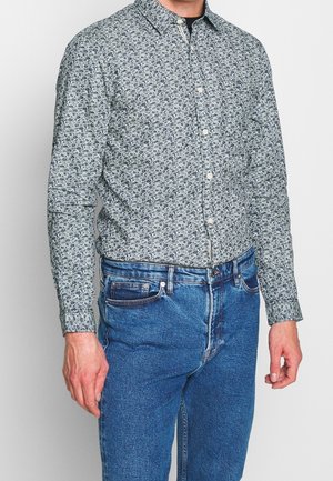 Homme portant une chemise à manches longues bleue à motifs floraux rentrée dans un jean bleu taille haute, se tenant contre un fond uni.