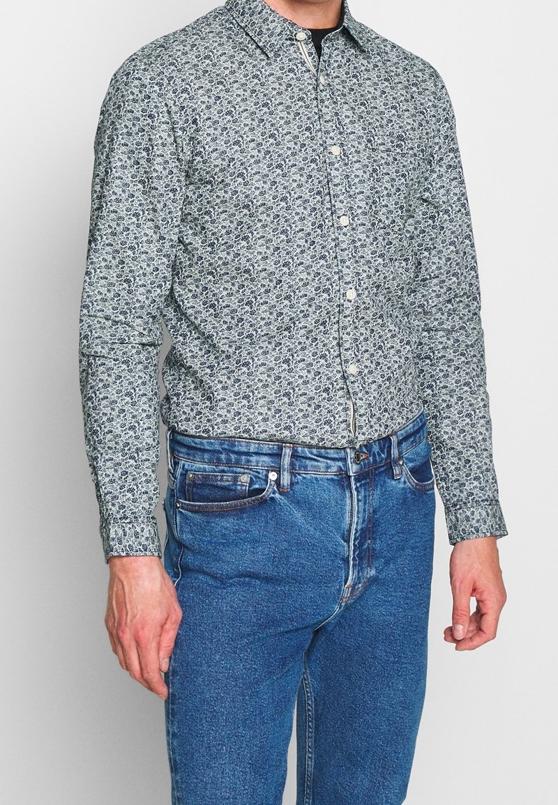 Man som bär en långärmad skjorta med blå blommönster, instoppad i högmidjade blå jeans, som står mot en enfärgad bakgrund.