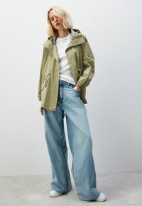 Jachetă verde măsliniu deschis, cu glugă, șnururi reglabile și buzunare laterale, asortată cu pantaloni din denim albastru deschis cu picioare largi și adidași albi.