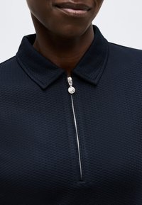 Κοντινό πλάνο ενός ατόμου που φοράει μπλε σκούρο πικέ πόλο με φερμουάρ και ασημένιο στρογγυλό τρέιλερ φερμουάρ.