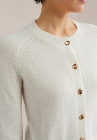 Stickad kofta i ljus kräm, med rundad halsringning, knappar i guldton och en mjuk textur med ribbade detaljer vid kragen och ärmsluten.