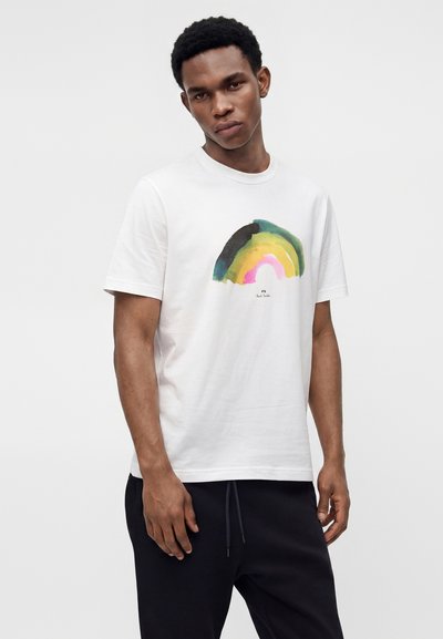Άνδρας που φοράει λευκό κοντομάνικο T-shirt με αφηρημένο ακουαρέλα ουράνιο τόξο σχεδιασμό, συνδυασμένο με μαύρο παντελόνι, στέκεται μπροστά σε απλό φόντο.