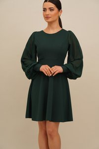 Kvinne iført en mørkegrønn knelang kjole med gjennomsiktige, voluminøse, lange ermer, stående med hendene foldet, iført gulløredobber og ringer.