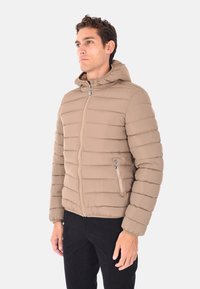 Uomo che indossa un giubbotto beige trapuntato con cappuccio e tasche con zip, e pantaloni neri, in piedi di fronte a uno sfondo bianco uniforme.